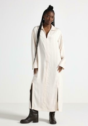 Donna con capelli lunghi intrecciati che indossa un vestito color crema, a maniche lunghe, lungo fino alla caviglia con spacco laterale e stivali neri fino a metà polpaccio.