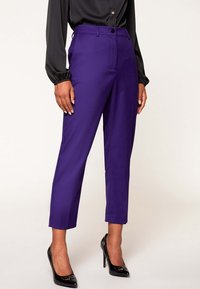 Lila ankellånga byxor med smal midja, knapp framtill och sidofickor, matchade med svarta högklackade skor och en blus.