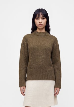 Jeune femme aux cheveux raides et foncés portant un pull en maille vert olive et une jupe côtelée de couleur crème, debout devant un fond uni.