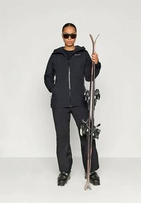 Skidkläder som inkluderar en svart vattentät jacka och byxor, håller skidor med bindningar, kompletterat med mörka solglasögon.