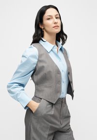 Γκρι καλοραμμένο γιλέκο πάνω από ένα ανοιχτόγαλάζιο πουκάμισο με κουμπιά. Το γιλέκο έχει χαμηλή λαιμόκοψη και δύο κουμπιά, με υφή υφάσματος και μπροστινές τσέπες.