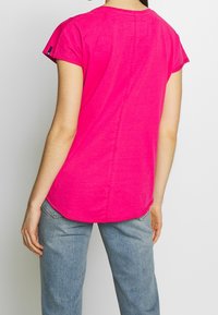 Persona vestida con una camiseta corta de color rosa brillante y jeans azul claro, de pie mirando hacia afuera contra un fondo simple.