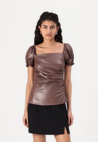 Haut marron, brillant, à manches courtes avec un décolleté carré, des manches bouffantes froncées et un ruché sur le devant. Porté avec une jupe noire.