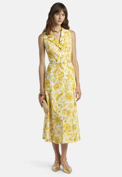 Kvinde iført gul og hvid blomsterprikket midi-kjole uden ærmer, guldkæde, armbånd, der holder en halmclutch, og gyldne spidse sko.