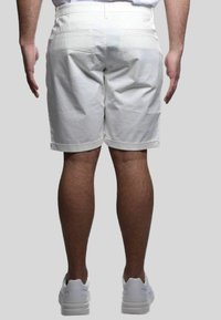 Shorts bianchi di cotone con vita piatta, due tasche posteriori e orlo arrotolato. Indossati con scarpe da ginnastica bianche, vista da dietro.