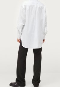 White long-sleeve shirt with a back yoke, curved hem, and side slits, paired with black wide-leg pants and black shoes.