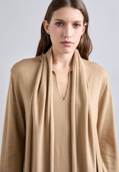 Lang beige cardigan med V-hals og en blød, tekstureret finish. Den har et matchende tørklæde draperet om halsen.