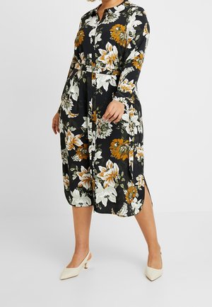 Femme portant une robe midi noire avec un grand imprimé floral blanc et orange, manches longues, fente latérale, et chaussures blanches à talons bas.