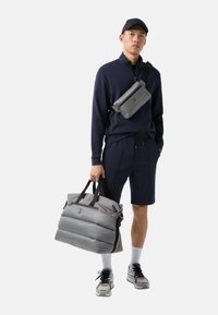 Pánský outfit zahrnující tmavě modrý pulovr, odpovídající kraťasy, šedou crossbody tašku a polstrovanou šedou cestovní tašku. Doplňky zahrnují bílé ponožky a tenisky.