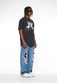 Schwarzes Grafik-T-Shirt mit einem blauen Vogeldesign, kombiniert mit hellblauen, stark zerrissenen Jeans. Weiße Sneakers runden den Look ab.