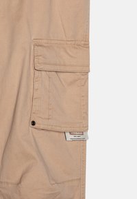 Beige cargo pants feature a side pocket with a flap, visible stitching, and a branded label near the hem. Fabric is smooth and durable.