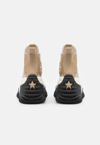 Sneakers typu high-top z błyszczącą beżową cholewką, czarną teksturowaną podeszwą i wyraźnymi akcentami w kształcie gwiazdy na pięcie. Wzmocnione szwy.