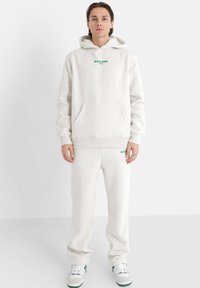 Conjunto de sudadera y pantalones de chándal en un material suave, similar a forro polar. Presenta un bolsillo frontal y texto de logo verde en el pecho y los pantalones.