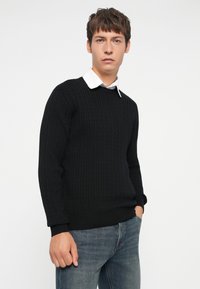 Pulover negru tricotat cu un model texturat, guler rotund și manșete cu dungi, purtat cu o cămașă albă cu guler dedesubt, asortat cu blugi albaștri.