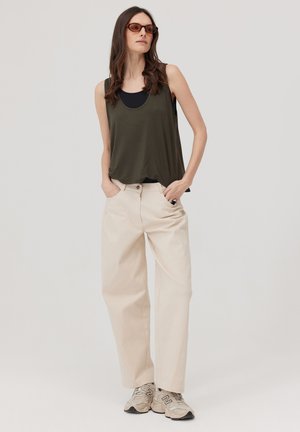 Kobieta w oliwkowym, bezrękawnym topie, beżowych szerokich spodniach, beżowych trampkach i brązowych okularach przeciwsłonecznych, stojąca z rękami w kieszeniach.