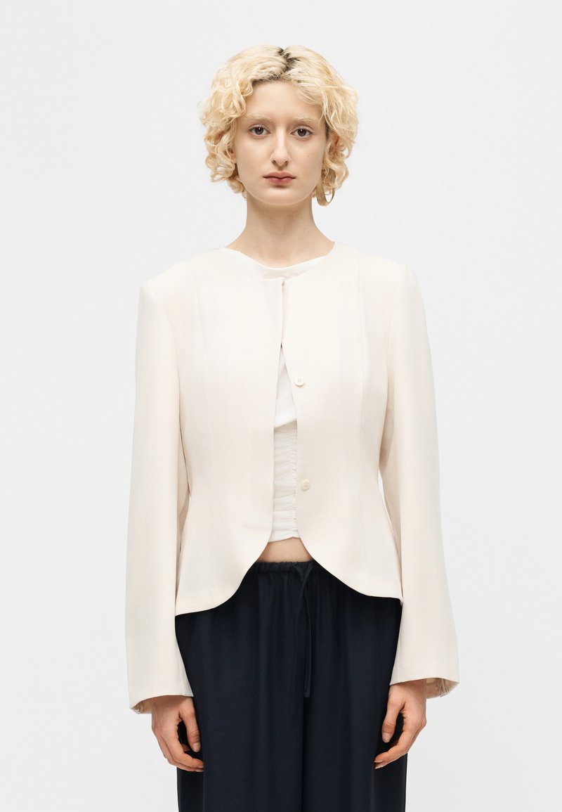 Άτομο με κοντά σγουρά ξανθά μαλλιά φοράει λευκό cropped σακάκι πάνω από λευκό τοπ και φαρδιά μαύρη παντελόνια πάνω σε απλό λευκό φόντο.