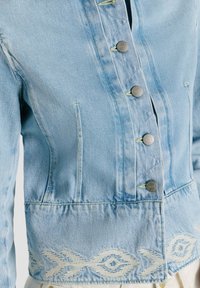 Giacca di jeans azzurro con bottoni in metallo e motivi geometrici ricamati bianchi lungo il bordo inferiore.