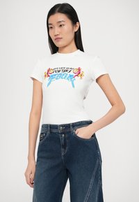 Biele priliehavé tričko s farebným grafickým dizajnom a textom "Versace Jeans Couture." Model má na sebe tmavé rifľové nohavice s prednými vreckami.