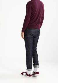 Mężczyzna w ciemnych dżinsach podwiniętych na mankietach, białych skarpetkach, burgundowych trampkach z białymi paskami i burgundowym swetrze z długim rękawem, stojący w profilu.