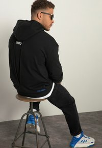 Felpa nera con la scritta "UNHOLY" sulla schiena, abbinata a jeans neri e sneakers blu, seduto su uno sgabello davanti a uno sfondo neutro.