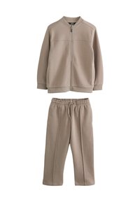 Veste beige zippée avec poches latérales et pantalon à taille élastique assorti. Les deux sont fabriqués en tissu doux avec une texture lisse. Design simple.