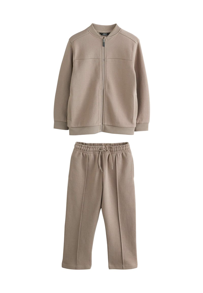 Veste beige zippée avec poches latérales et pantalon à taille élastique assorti. Les deux sont fabriqués en tissu doux avec une texture lisse. Design simple.