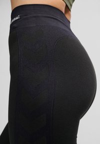 Nærbillede af en person iført sort højttaljede leggings med diskrete chevronmønstre og et lille logo nær taljebåndet.