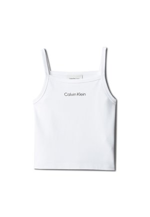 Ragazza con lunghi capelli castani che indossa una canotta bianca Calvin Klein, gonna di jeans chiara, sandali beige, in piedi con le mani nelle tasche.