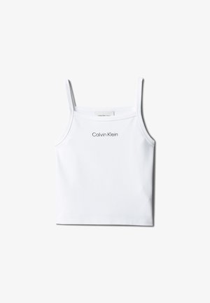 Dziewczyna o długich brązowych włosach, ubrana w biały top Calvin Klein, jasną dżinsową spódnicę, beżowe sandały, stojąca z rękami w kieszeniach.