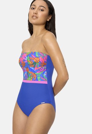 Mujer con traje de baño azul sin tirantes con la parte superior estampada con flores y una banda rosa en la cintura, parada contra un fondo claro y sencillo.