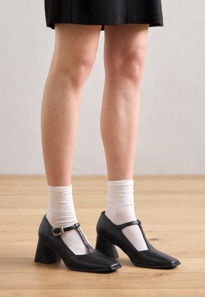 Zwarte Mary Jane-hakken met een vierkante neus, T-band en gespsluiting. Gecombineerd met witte enkelsokken op een houten vloer.