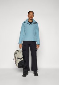 Giacca impermeabile azzurro chiaro con cappuccio, maglietta nera sottostante, pantaloni cargo e stivaletti neri robusti; tiene uno zaino grigio con dettagli neri.