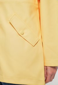 Μανίκι κίτρινου αδιάβροχου μπουφάν και καπάκι τσέπης με κουμπιά, με μέρος ενός χεριού και μπλε τζιν παντελόνι ορατό.