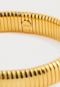 Gold-tone circular bracelet with tightly spaced ridges, featuring a small round charm engraved with "Luvaj." Smooth, shiny surface.
