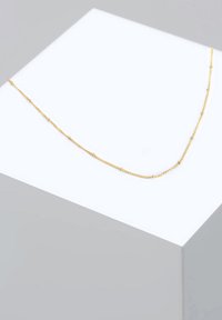 Collier couleur or avec une chaîne délicate, comportant de petites accents ronds uniformément espacés le long de sa longueur, sur un support d'exposition blanc.