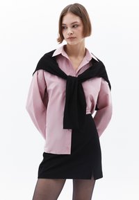 Lichtroze button-up shirt met lange mouwen, zwarte trui geknoopt om de nek en zwarte mini-jurk met een zijsplit, gedragen met donkere panty's.