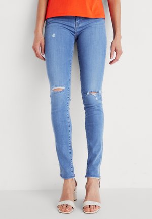 Femme portant un jean skinny bleu clair déchiré, un haut orange et des sandales à talons blocs blanches à bout ouvert sur fond blanc uni.