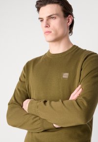 Oliwkowy sweter z dzianiny z okrągłym dekoltem i ściągaczami, z kwadratową naszywką logo na klatce piersiowej. Miękka tekstura i dopasowany krój.