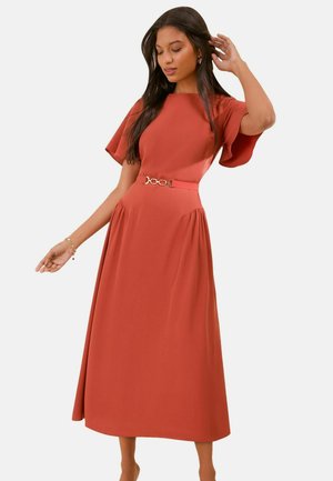 Femeie purtând o rochie midi de culoare ruginiu cu mâneci fluture și o curea subțire, pozând cu o mână lângă cap și cealaltă coborâtă.