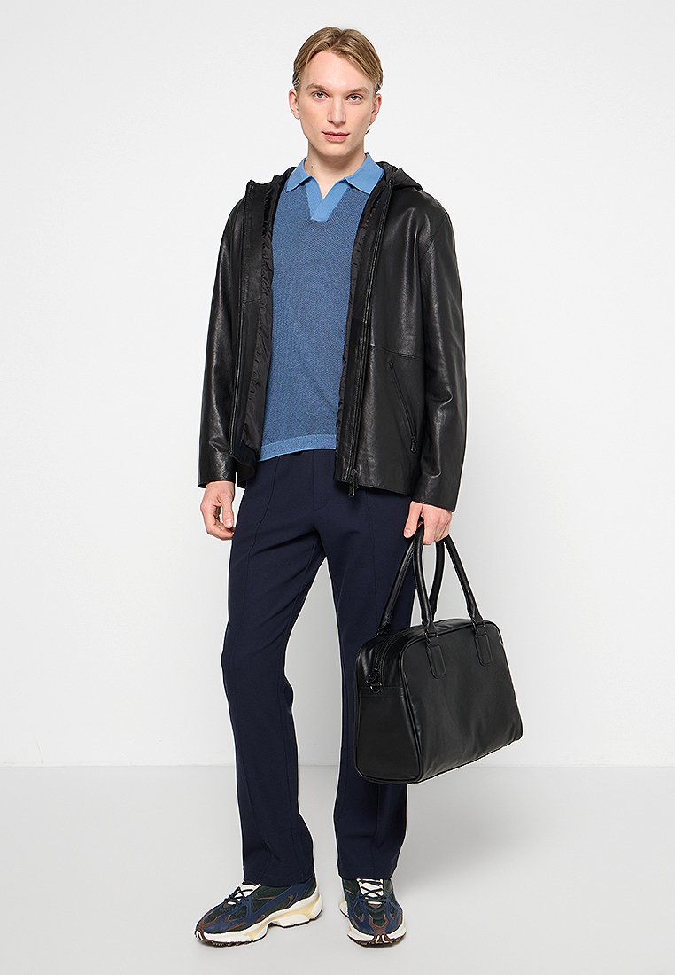 Schwarze Lederjacke über einem blauen Pullover, marineblaue Hose und Sport Schuhe. Trägt eine schwarze Lederhandtasche mit doppelten Griffen.