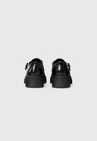 Pair of black leather platform shoes with gold buckle accents, shown from the back on a plain light gray background.