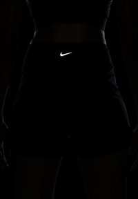 Atleta vistiendo un pantalón corto negro de Nike y un sujetador deportivo, de pie con los brazos relajados a los lados contra un fondo oscuro.