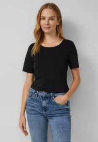 Schwarzes Kurzarm-T-Shirt mit rundem Ausschnitt, aus weichem Stoff, kombiniert mit mittelblauen Jeans, die Hände in den Taschen.