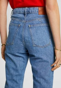 Hochgeschnittene blaue Jeans aus Denim mit strukturiertem Finish, ausgestattet mit Gesäßtaschen und einem Lederetikett. Kombiniert mit einem roten Oberteil und goldenen Accessoires.