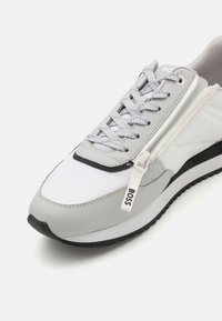 Harmaat ja valkoiset urheilukengät siistillä muotoilulla, joissa on vetoketjusomiste, teksturoidut materiaalit ja musta kumipohja klassisella kuvioinnilla.