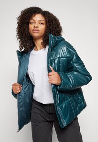 Tyrkysová nafukovacia bunda s lesklou prešívanou textúrou, zipsom vpredu a nastaviteľnou kapucňou, nosená cez biely tričko a tmavé džínsy.