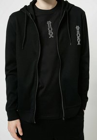 Felpa nera con zip e coulisse, con una grafica bianca a contrasto sul petto. Tessuto morbido, polsini fitted e tasca a marsupio.