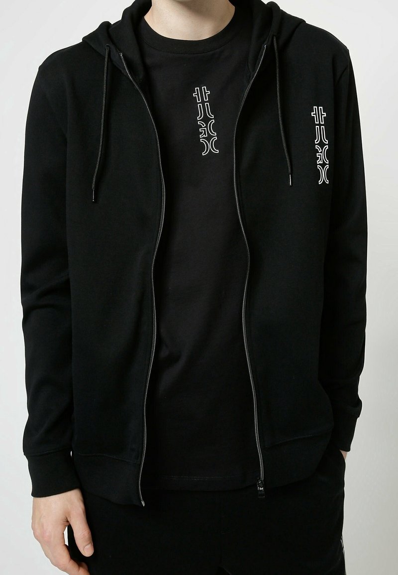 Felpa nera con zip e coulisse, con una grafica bianca a contrasto sul petto. Tessuto morbido, polsini fitted e tasca a marsupio.