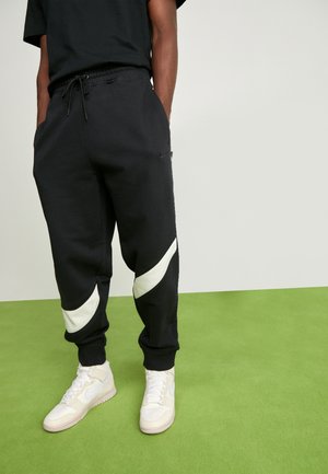 Osoba nosząca czarne dresy z dużym białym logo Nike na nogawkach oraz białe sneakersy Nike, stojąca na zielonej podłodze.