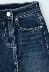 Jean en denim bleu foncé en gros plan montrant une poche avant, un bouton avec logo, des rivets métalliques et des coutures beiges sur fond clair.